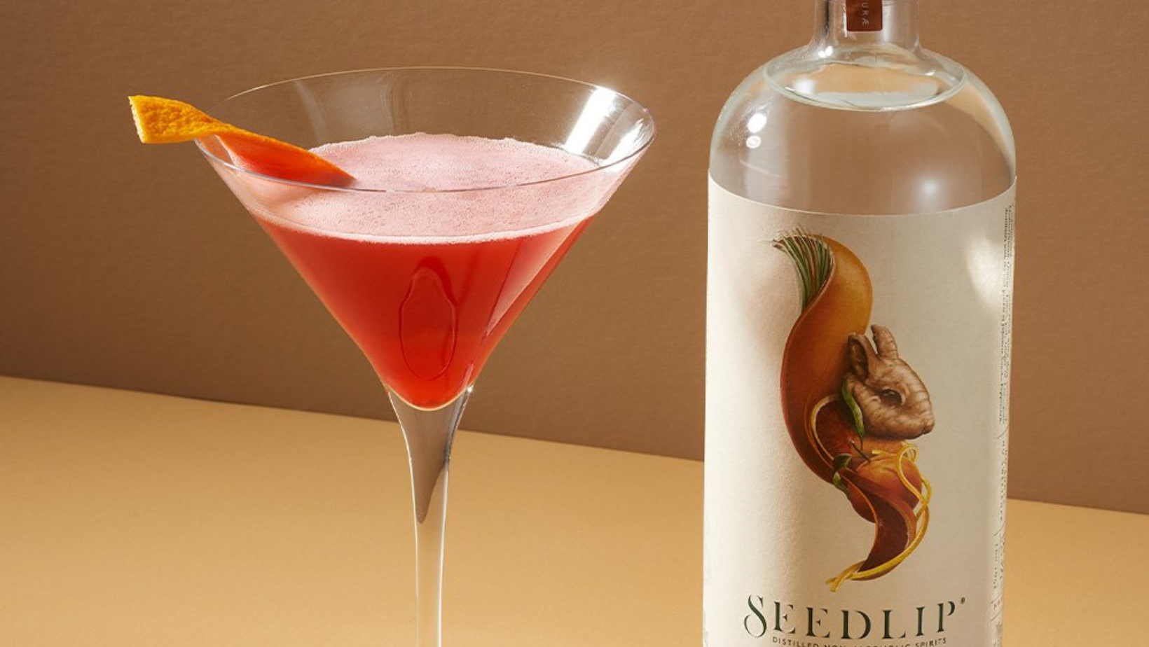 Cocktail Cosmopolitan Seedlip servi dans une coupe élégante, garni d’un zeste d’orange, avec une bouteille de Seedlip Grove 42 en arrière-plan | MAISON COCKTAIL